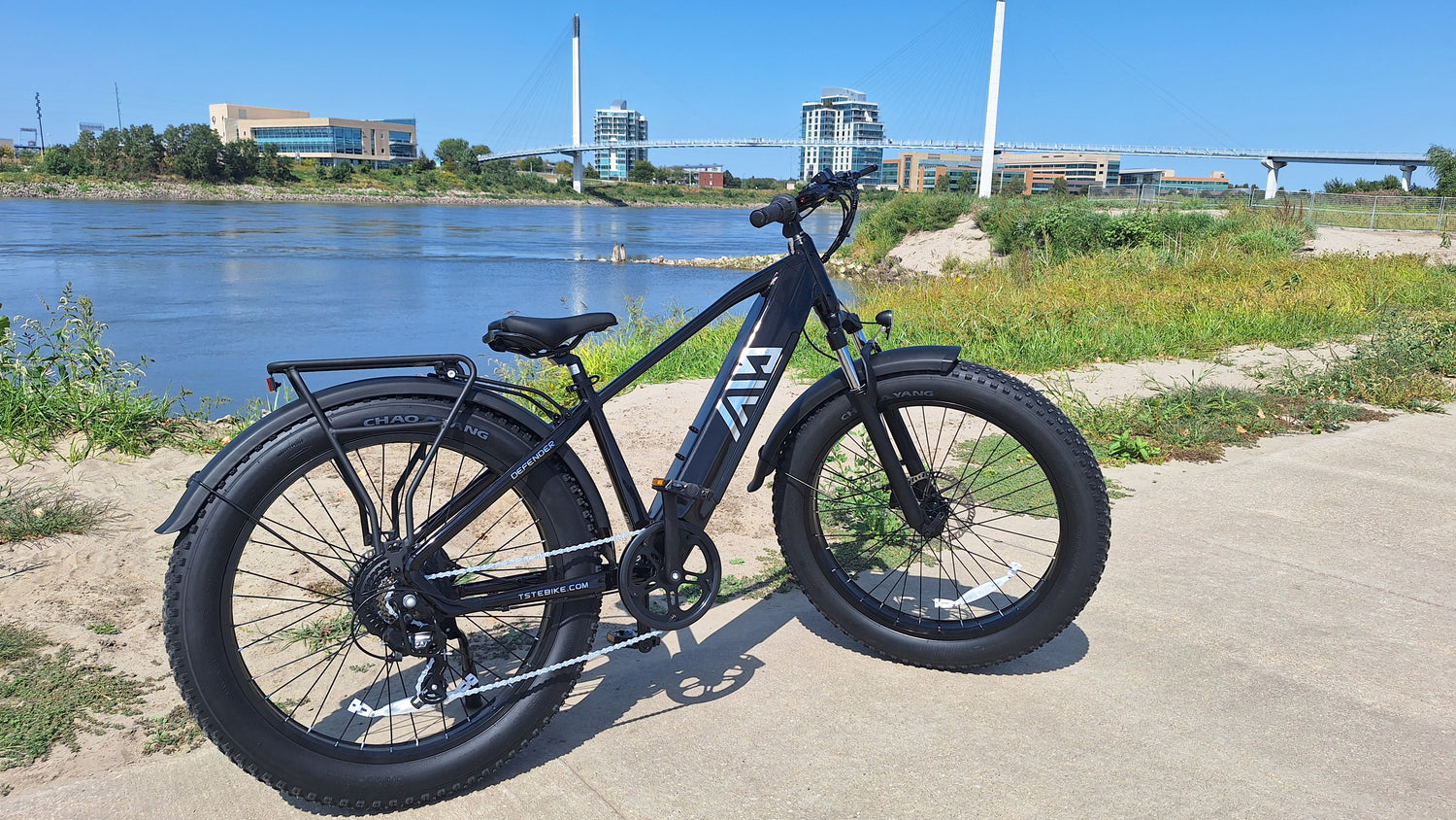 A TST Defender fat-tire electric bike is parked on a paved path next to a river with a modern cityscape and bridge in the background.